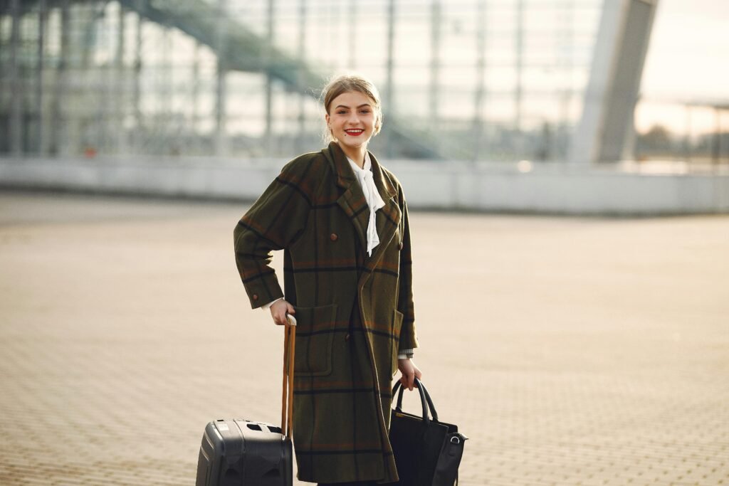 pexels photo 3876042 3876042 Positive smiling young female traveler in casual warm coat with suitcase and ladies bag standing near airport terminal and looking at camera