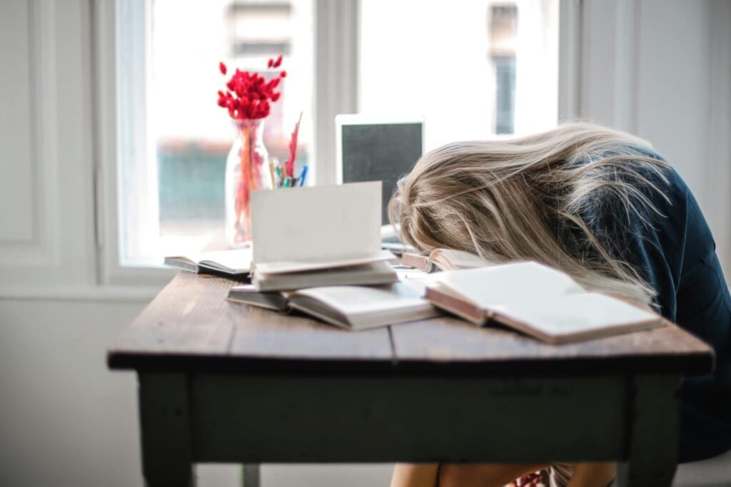 Blonde woman slumped over desk filled with books, showcasing study fatigue indoors. (Burnout)